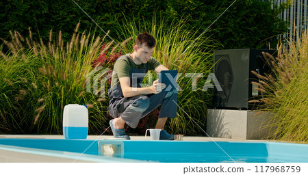 Pool technician using a tablet by the pool, surrounded by greenery and a chemical container, focused on maintenance tasks. Pool technician using a tablet by the pool, surrounded by greenery and a chemical container, focused on maintenance tasks. 117968759