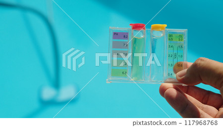 A hand holds a water test kit over a bright blue pool, with sunlight reflecting off the water's surface. 117968768