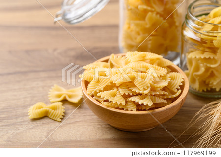 Uncooked farfalle pasta (bow tie shape) in wooden bowl and glass jar, Italian food ingredient Uncooked farfalle pasta (bow tie shape) in wooden bowl and glass jar, Italian food ingredient 117969199
