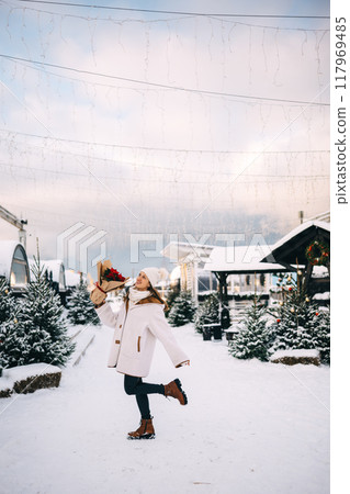 A woman joyfully poses in a winter wonderland while holding flowers amidst snowy pines A woman joyfully poses in a winter wonderland while holding flowers amidst snowy pines 117969485