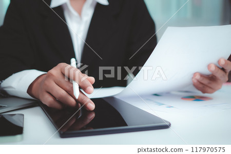Businesswoman in black suit working on laptop computer, paper documents, tablet, and calculator, Hands touch on tablet at office with dark background, Online working, Close up, Copy space. Businesswoman in black suit working on laptop computer, paper documents, tablet, and calculator, Hands touch on tablet at office with dark background, Online working, Close up, Copy space. 117970575