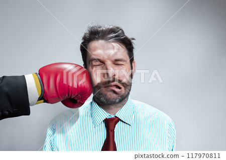 Man in business suit takes punch to face from red boxing glove, his expression frozen in shock and impact, symbolizing tough business blow. 117970811