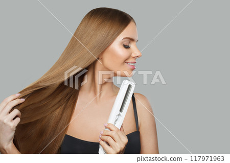 Attractive brunette woman holding straight hair iron and straightening her long smooth shiny hair  117971963