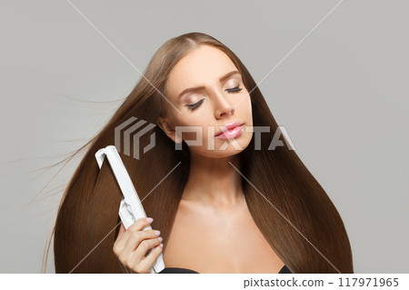 Pretty brunette woman holding straight hair iron and straightening her long smooth shiny hair Pretty brunette woman holding straight hair iron and straightening her long smooth shiny hair 117971965