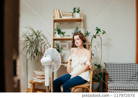 fan cools unhappy Caucasian woman languishing in summer heat. warmed-up experiences hot flashes 117972078