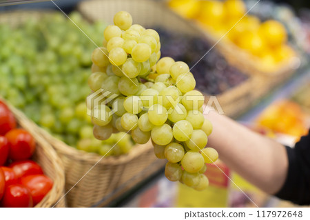 Hands hold a bunch of green grapes against the background of a blurred store counter. 117972648