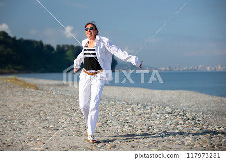 A middle-aged woman in white clothes walks along a pebble windy beach. A middle-aged woman in white clothes walks along a pebble windy beach. 117973281