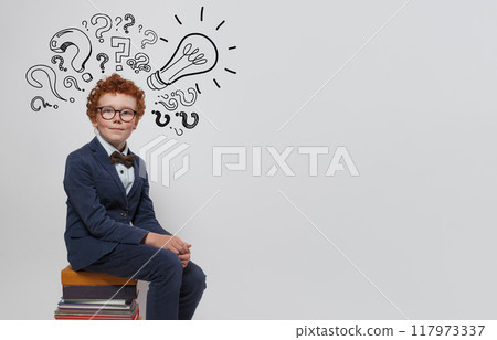 Cute child boy in guggles with light bulb and question marks sitting on stack of books Cute child boy in guggles with light bulb and question marks sitting on stack of books 117973337