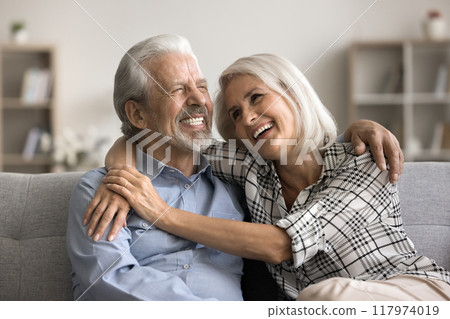 Cheerful attractive wellbeing mature spouses embracing sitting on couch Cheerful attractive wellbeing mature spouses embracing sitting on couch 117974019