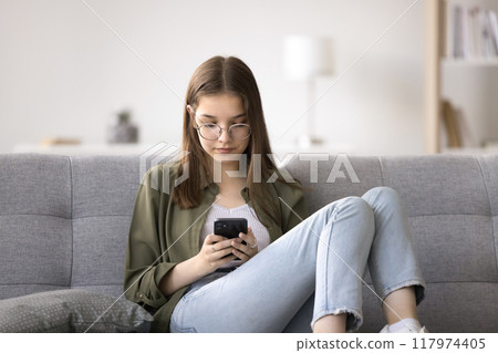 Pretty teen girl in glasses sitting on couch with smartphone Pretty teen girl in glasses sitting on couch with smartphone 117974405
