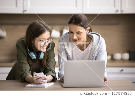 Teen girl and mother using smartphone and laptop in kitchen Teen girl and mother using smartphone and laptop in kitchen 117974533