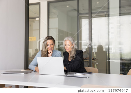 Positive younger and older businesswomen watching online content on laptop Positive younger and older businesswomen watching online content on laptop 117974947