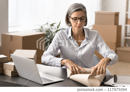 Serious mature woman wrapping parcel with paper Serious mature woman wrapping parcel with paper 117975094