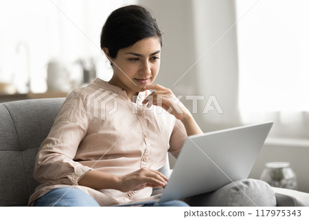 Pensive Indian woman learn program studying on-line seated on sofa Pensive Indian woman learn program studying on-line seated on sofa 117975343