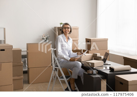 Middle-aged woman managing tasks in a workspace filled with boxes Middle-aged woman managing tasks in a workspace filled with boxes 117975406