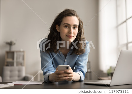 Pretty woman leans at desk holding smartphone looking at camera 117975511