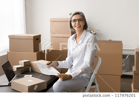 Mature woman sits in workspace surrounded by boxes and packages Mature woman sits in workspace surrounded by boxes and packages 117976201