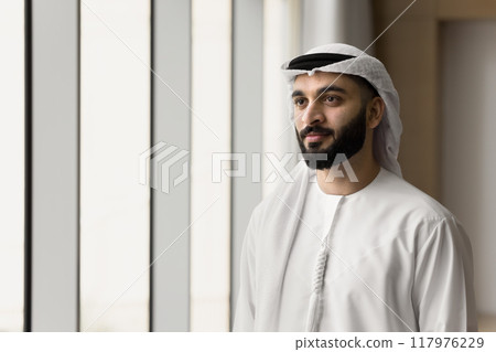 Positive handsome young Arabic businessman standing at office window Positive handsome young Arabic businessman standing at office window 117976229