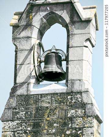 A bell is mounted on a stone tower. The bell is made of metal and has a circular frame. The tower is made of light-colored stone with visible wear and tear. 117977205