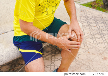A middle-aged man who is jogging is sitting on a bench and putting his hands on his knees to endure the pain in his knees A middle-aged man who is jogging is sitting on a bench and putting his hands on his knees to endure the pain in his knees 117977780