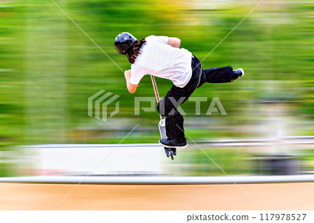 Young man practicing Scootering (Freestyle Scootering) in the new SkatePark in the central park of Igualada, Barcelona, Spain. blurred background Young man practicing Scootering (Freestyle Scootering) in the new SkatePark in the central park of Igualada, Barcelona, Spain. blurred background 117978527