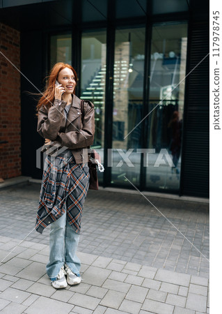 Vertical portrait of pretty young woman with long red hair and freckles smiling chatting on smartphone outdoors. Cute redhead female dressed in leather jacket, enjoying conversation in urban setting Vertical portrait of pretty young woman with long red hair and freckles smiling chatting on smartphone outdoors. Cute redhead female dressed in leather jacket, enjoying conversation in urban setting 117978745