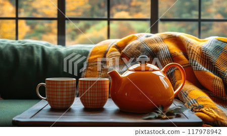 A vibrant orange teapot sits on a wooden tray accompanied by two checkered mugs, surrounded by a cozy blanket, in a serene autumn setting by the window. 117978942