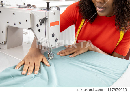 African American woman sewing clothes from light blue textile with sewing machine African American woman sewing clothes from light blue textile with sewing machine 117979013