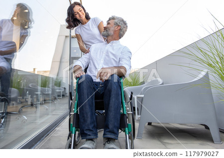 Dark-haired woman carrying a wheelchair of her husband 117979027