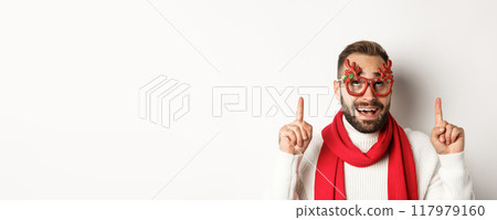 Christmas, New Year and celebration concept. Close-up of excited bearded guy in party glasses, pointing and looking up at store promo, white background 117979160