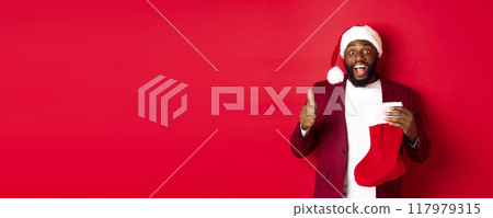 Excited Black man showing thumb-up in approval, holding christmas sock with holiday gifts, smiling amazed, standing over red background 117979315