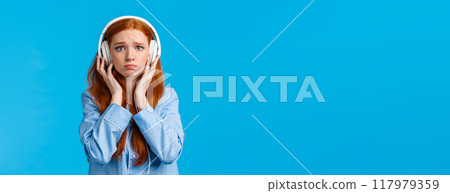 Girl about cry from emotional song. Upset redhead girlfriend suffering breakup, sobbing listening music in large white headphones, standing blue background in nightwear, unhappy Girl about cry from emotional song. Upset redhead girlfriend suffering breakup, sobbing listening music in large white headphones, standing blue background in nightwear, unhappy 117979359
