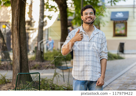 Happy young Indian man looking approvingly at camera showing thumbs up like gesture on city street Happy young Indian man looking approvingly at camera showing thumbs up like gesture on city street 117979422