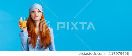 Girl likes drinking fresh juices. Cheerful and cute pretty feminine ginger girl, foxy teenager in nightwear, sleep mask, biting lip eager, seeing something tempting, holding glass orange juice 117979498