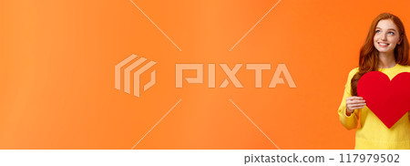 Dreamy cute redhead girl waiting for her prince, planning perfect velantines day romantic date, holding large heart sign and looking upper left corner with tender happy smile, orange background Dreamy cute redhead girl waiting for her prince, planning perfect velantines day romantic date, holding large heart sign and looking upper left corner with tender happy smile, orange background 117979502