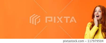 Fascinated and amiring cute redhead woman looking impressed and excited at upper left corner, smiling see breathtaking view, cover opened mouth palm thrilled, losing speech happy, orange background 117979504