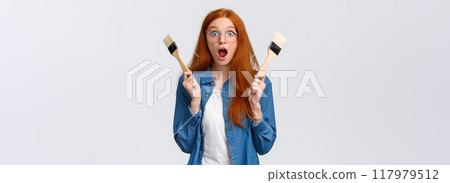 Amused and excited, wondered redhead woman creating new art, painting, look stunned and thrilled, say wow drop jaw and pop eyes camera, holding two paintbrushes, white background 117979512