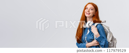 College life, modern lifestyle and education concept. Cheerful good-looking redhead female student with foxy long hair, wearing headphones over neck, backpack, smiling camera 117979529