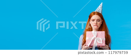 Tempting moment of unwrapping gift. Cute glamour redhead teenage girl biting lip eager open cute birthday present, wearing silly cap, celebrating b-day standing blue background curious Tempting moment of unwrapping gift. Cute glamour redhead teenage girl biting lip eager open cute birthday present, wearing silly cap, celebrating b-day standing blue background curious 117979739