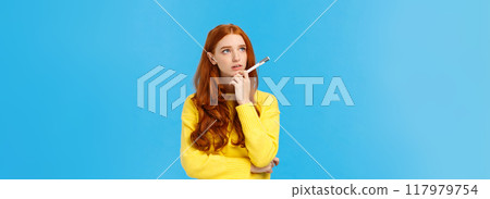 Troubled, hesitant and indecisive redhead, ginger girl with curly hair in yellow sweater, looking unsure upper left corner as pondering, solving exercise in mind, holding pen, searching inspiration 117979754