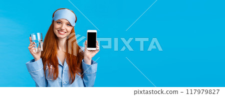 Waist-up portrait elegant and glamour redhead female in nightwear, sleep mask, holding glass water and smiling, holding smartphone, showing mobile app on screen, blue background Waist-up portrait elegant and glamour redhead female in nightwear, sleep mask, holding glass water and smiling, holding smartphone, showing mobile app on screen, blue background 117979827