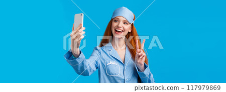 Cute outgoing redhead female want followers check out her new nightwear, extend hand with smartphone, taking selfie and smiling joyfuly make peace gesture express positivity, blue background Cute outgoing redhead female want followers check out her new nightwear, extend hand with smartphone, taking selfie and smiling joyfuly make peace gesture express positivity, blue background 117979869