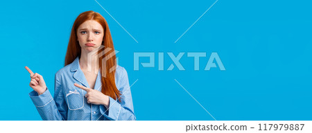 Regret, bad mood and advertising concept. Sad unhappy redhead girl in nightwear, sulking and frowning express disappointment or jealousy, pointing fingers right, standing blue background 117979887