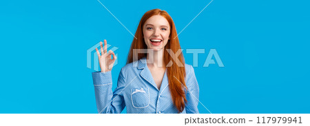 Alright, no worries anymore. Cheerful and relieved smiling happy redhead girl in nightwear, ready sleep showing okay gesture like or approval, give positive reply, standing blue background 117979941