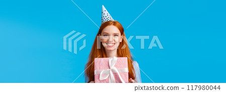 Waist-up portrait cheerful happy, pretty redhead, ginger girl celebrating birthday, receive cute pink wrapped present, wearing b-day cap and smiling, having fun at party, blue background 117980044