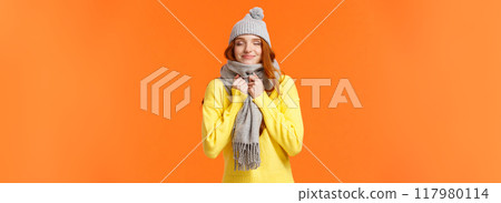 Cute and happy redhead woman walking along winter holidays fair market, close eyes and smiling as smelling something delicious, wearing warm grey hat and scarf, standing orange background Cute and happy redhead woman walking along winter holidays fair market, close eyes and smiling as smelling something delicious, wearing warm grey hat and scarf, standing orange background 117980114