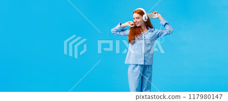 Full-length vertical shot cheerful and upbeat, carefree redhead teenage girl dancing and enjoying beautiful morning, wearing nightwear skipping college stay home listen music in headphones 117980147