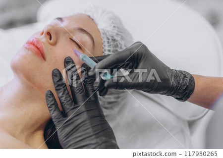 A professional beautician injects Botox into the face of a young woman 117980265