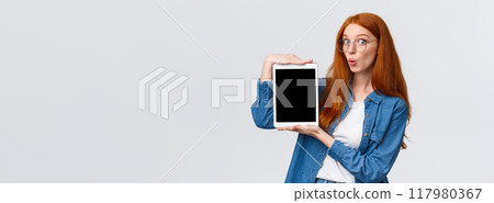 Amused, excited good-looking redhead woman in denim shirt, folding lips in wow sound, looking amazed camera, holding digital tablet, showing device screen, standing white background 117980367