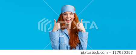 Waist-up portrait delighted, fully satisfied cute redhead girl in sleeping clothes, nightwear and sleep mask, showing thumbs-up in approval, like or agree gesture, standing blue background amused 117980397